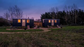 Two small cabins with windows, a picnic table, and a bench in a grassy area with trees in the background.