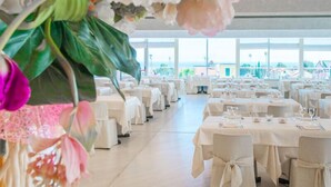 A spacious dining area with multiple tables set for guests, large windows offering a view of the sea, and a ceiling adorned with floral arrangements. - Residence Loano 2 Village  Studio Apartment 3 Pax (Loano)