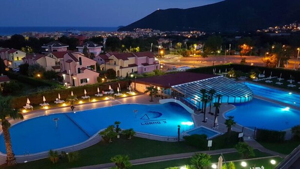 A swimming pool with a lounge area, surrounded by residential buildings and a view of the sea. - Residence Loano 2 Village  Studio Apartment 3 Pax (Loano)