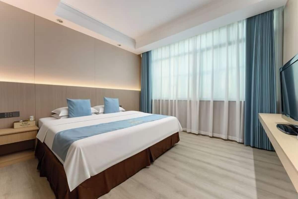 A hotel room with a bed, bedside table, TV, and large windows with curtains.