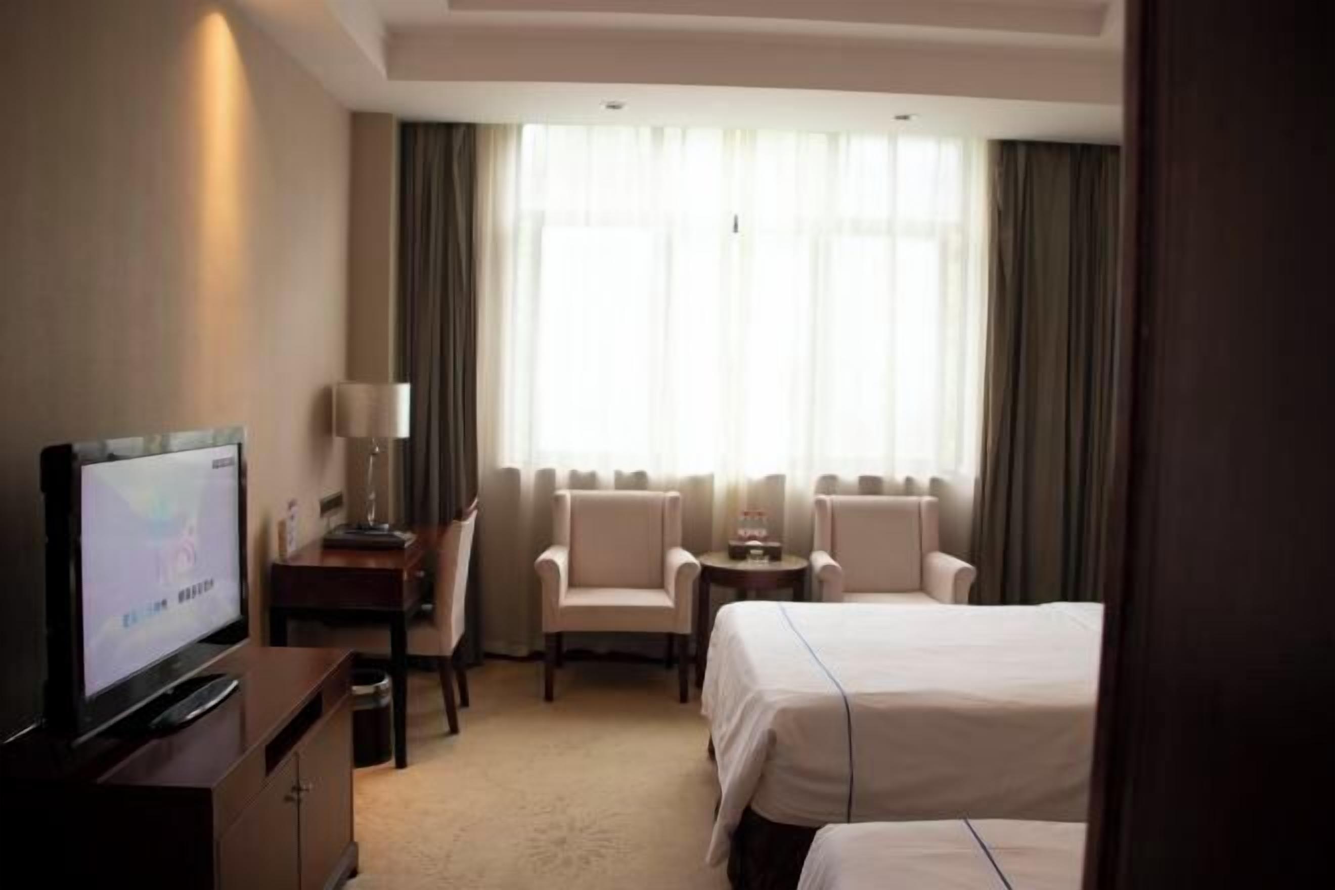 A hotel room with a bed, a desk with a television, two armchairs, a small table, and a large window with curtains.