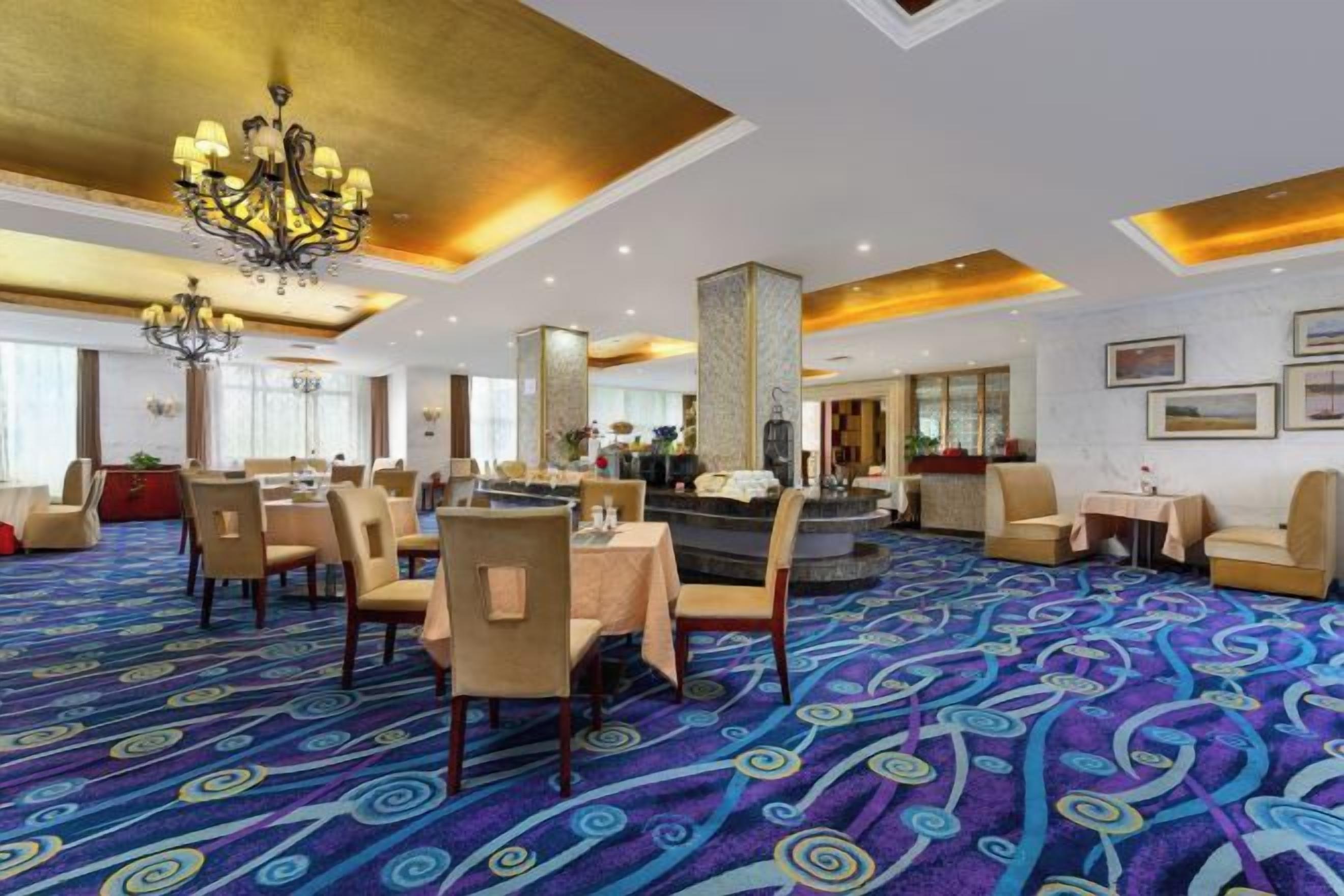 A hotel lobby with a patterned carpet, dining area with tables and chairs, a bar area, and a reception desk.