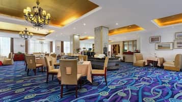 A hotel lobby with a patterned carpet, dining area with tables and chairs, a bar area, and a reception desk.