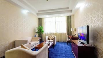 A hotel room with a large flat-screen TV, a sofa, a chair, a coffee table, and a potted plant.