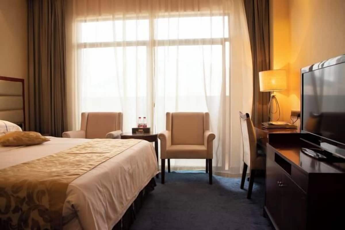 A hotel room with a bed, two armchairs, a desk with a lamp, a television, and a large window with curtains.