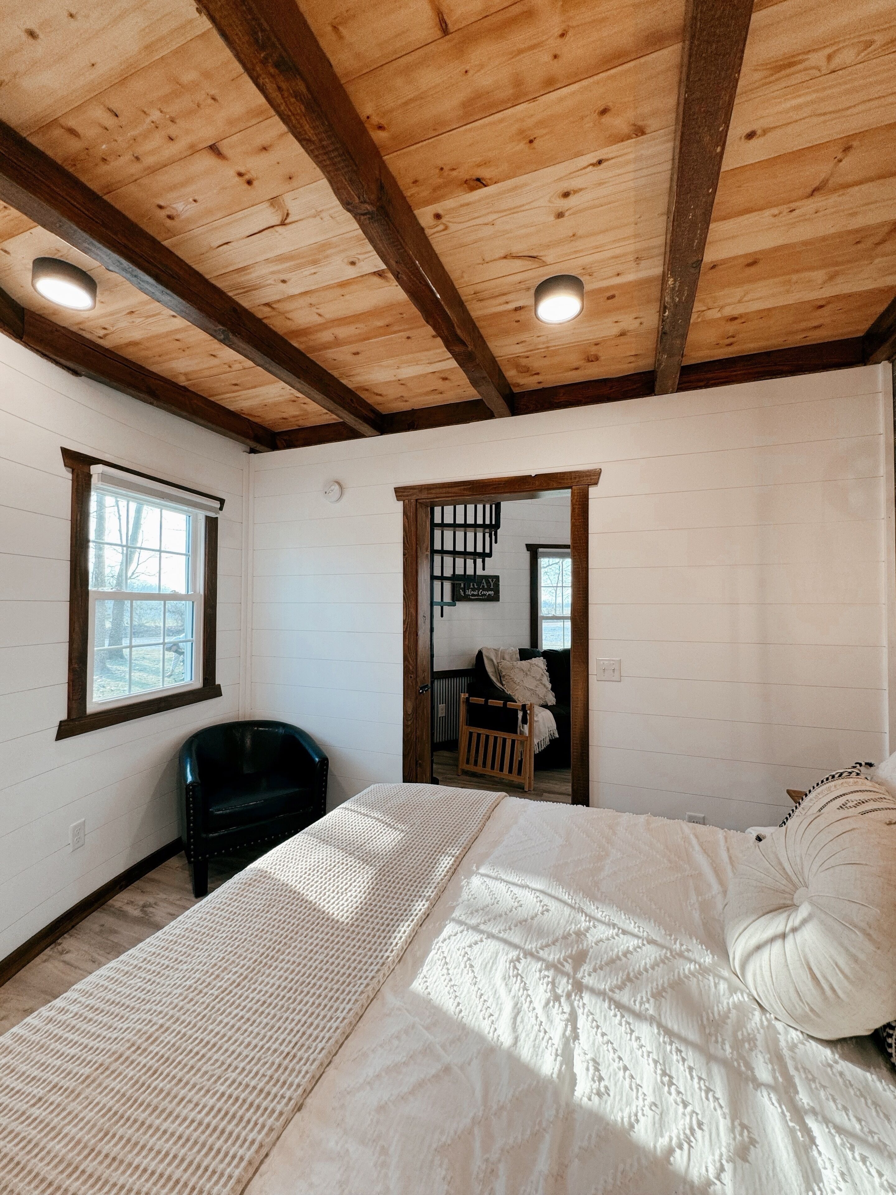 A bedroom with a wooden ceiling, a bed with white bedding, a black chair, and a window.
