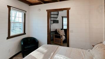 A bedroom with a wooden ceiling, a bed with white bedding, a black chair, and a window.