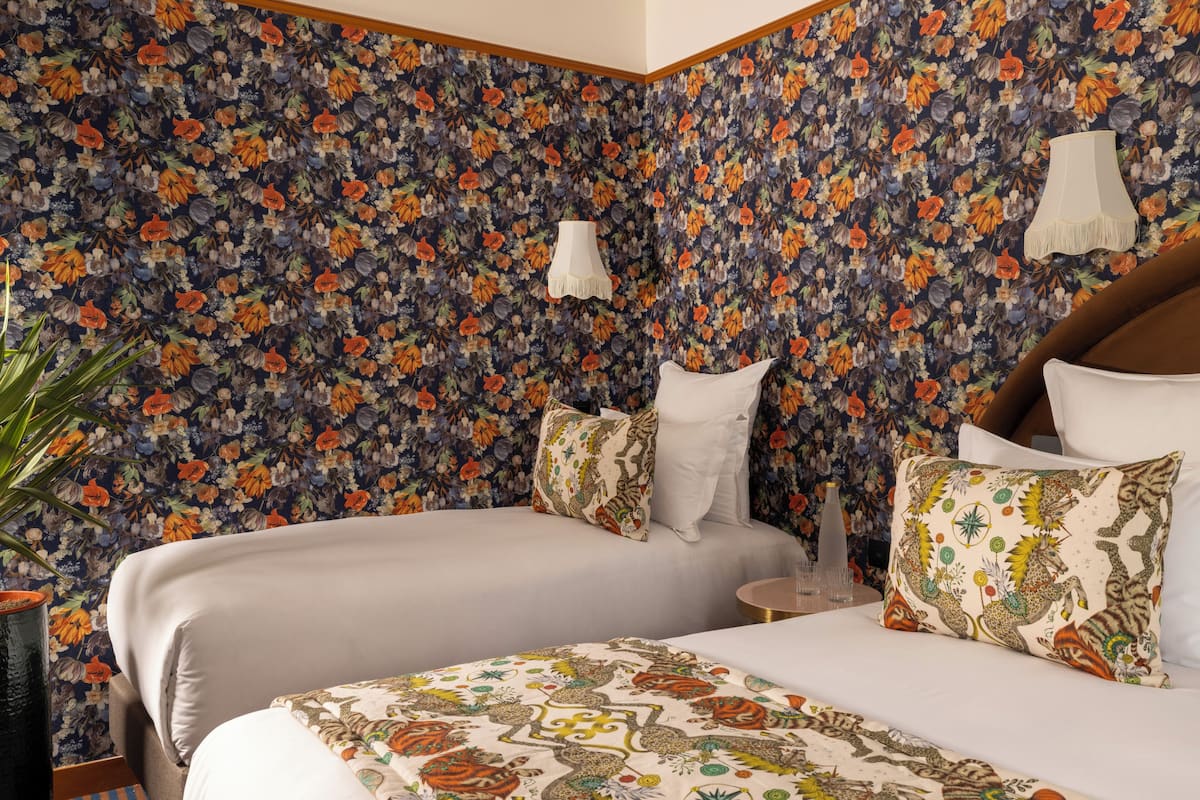 A hotel room with two beds, floral wallpaper, a potted plant, and a small table with a lamp.
