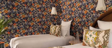 A hotel room with two beds, floral wallpaper, a potted plant, and a small table with a lamp.