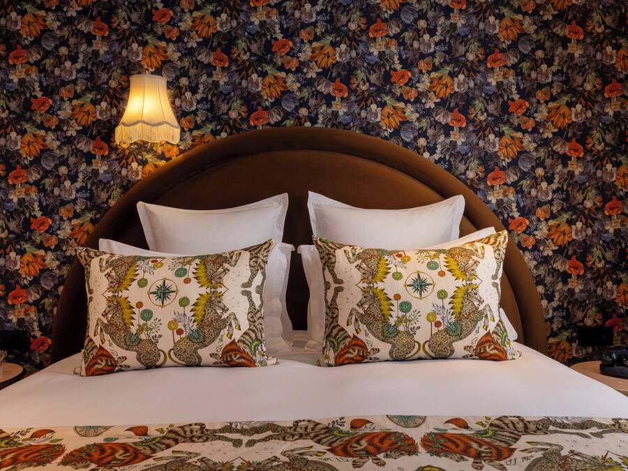 Un llit amb capçalera estampada amb flors, dos coixins decoratius, una làmpada de nit i paper pintat amb motius florals.