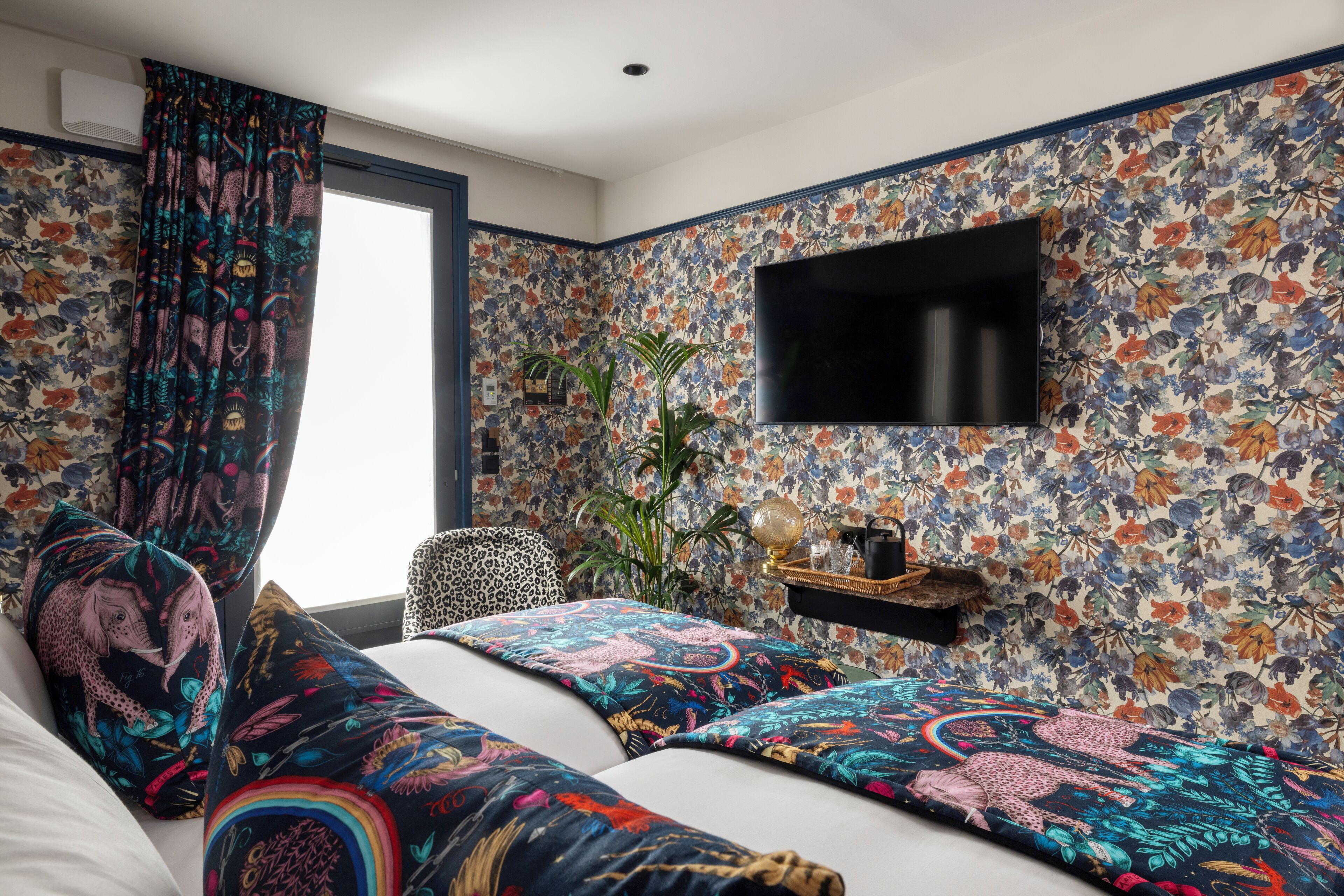 A bedroom with a floral wallpaper, a flat-screen TV mounted on the wall, a bed with colorful pillows, a plant, and a small shelf with items.