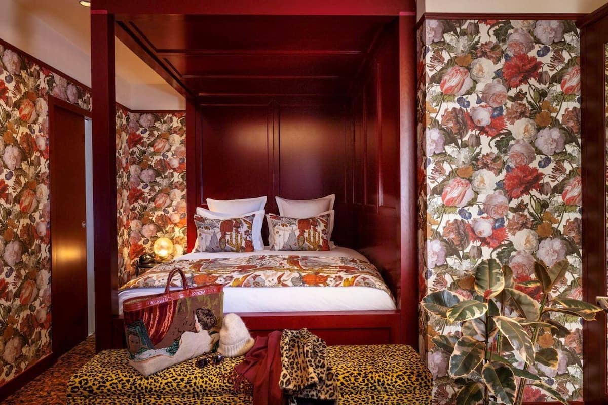 A bedroom with a four-poster bed, floral wallpaper, and a leopard print ottoman.