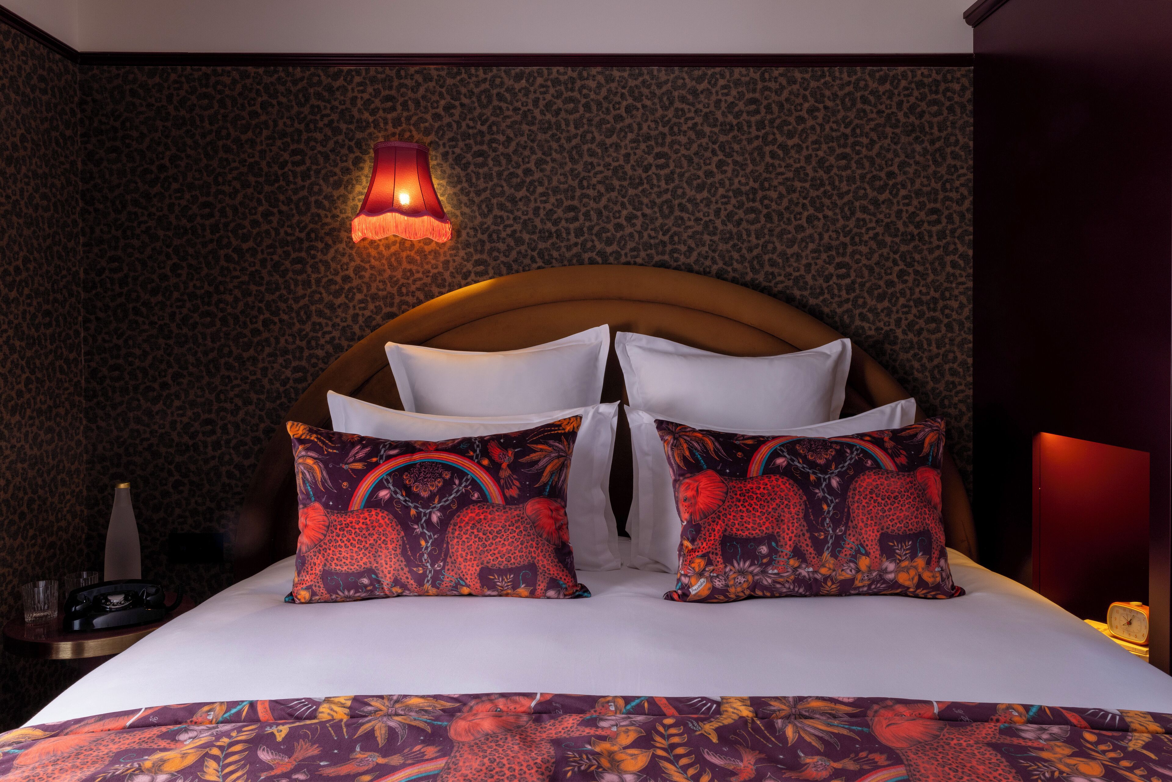 A hotel room with a leopard print wallpaper, a wooden headboard, white bedding, and colorful patterned pillows.