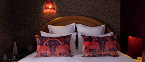 A hotel room with a leopard print wallpaper, a wooden headboard, white bedding, and colorful patterned pillows.