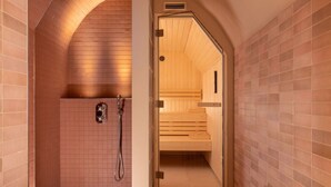 A sauna room with a wooden bench, a shower area with a handheld showerhead, and a tiled wall. - Ginette à la Folie (Paris)