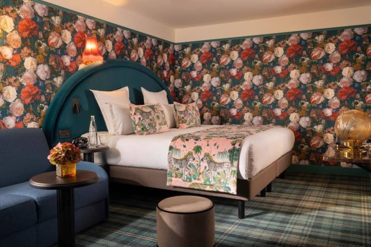 A bedroom with a floral wallpaper, a teal headboard, a bed with white bedding, a blue sofa, a small round table with a vase, and a plaid carpet.
