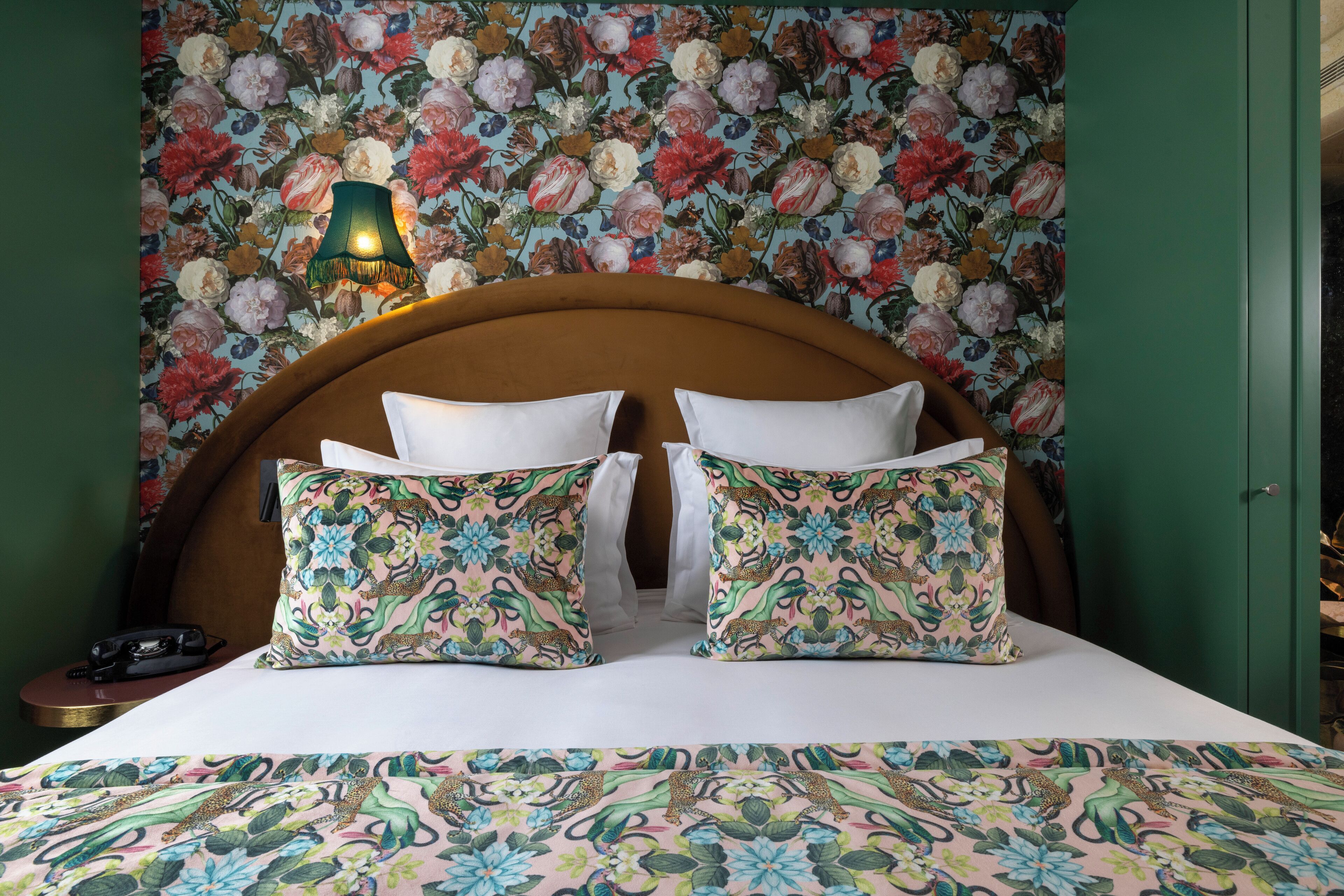Una cama con almohadas estampadas con motivos florales y una colcha a juego, una pared verde con papel tapiz floral, y una puerta verde.