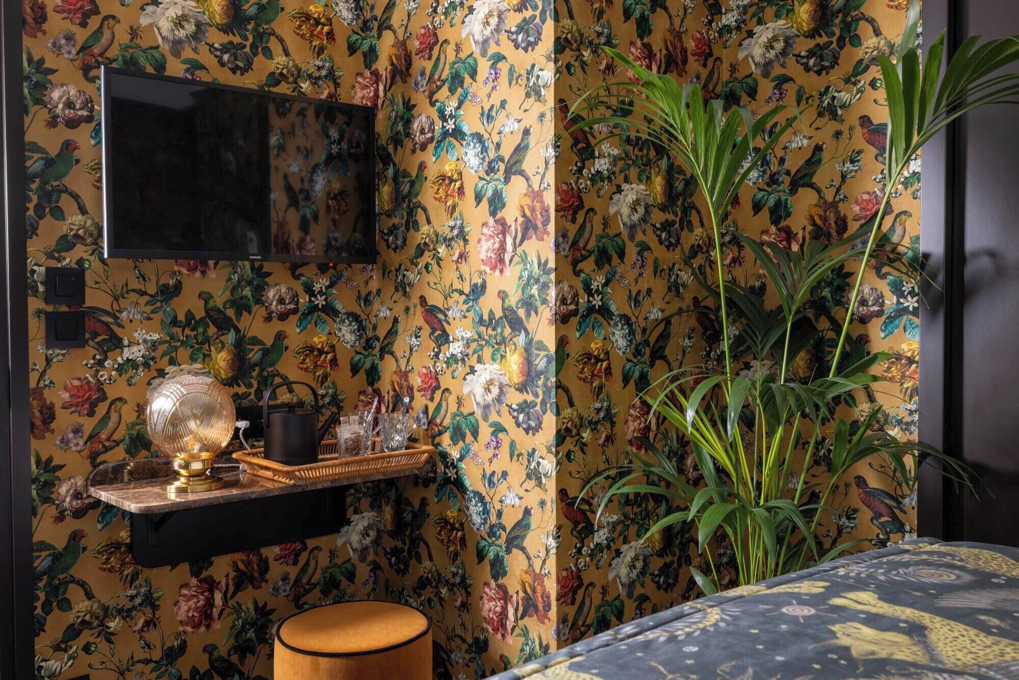 Een kamer met bloemenbehang, een aan de muur bevestigde televisie, een plant en een tafeltje met een lamp en decoratieve objecten.