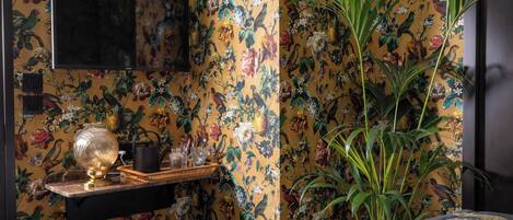 Quarto com papel de parede floral, televisão na parede, planta e uma mesa pequena com abajur e objetos decorativos.