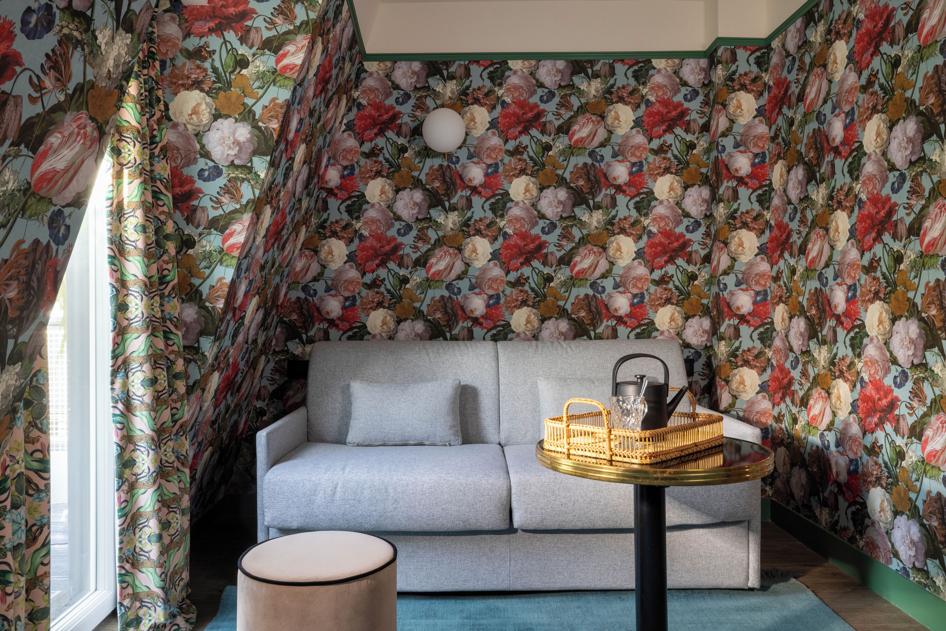 Um quarto com papel de parede floral, um sofá cinza, uma mesa redonda pequena e uma janela com cortinas.