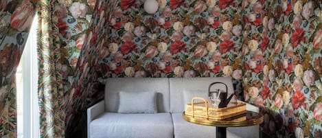 Um quarto com papel de parede floral, um sofá cinza, uma mesa redonda pequena e uma janela com cortinas.