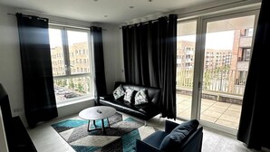 A modern living room with a black leather sofa, a blue armchair, and a round coffee table. There is a large window with black curtains, and a view of a residential area outside. - Frogley Park Cozy Apartment (Barking)