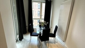 A modern dining area with a glass table and black chairs, a window with curtains, and a radiator on the wall. - Frogley Park Cozy Apartment (Barking)