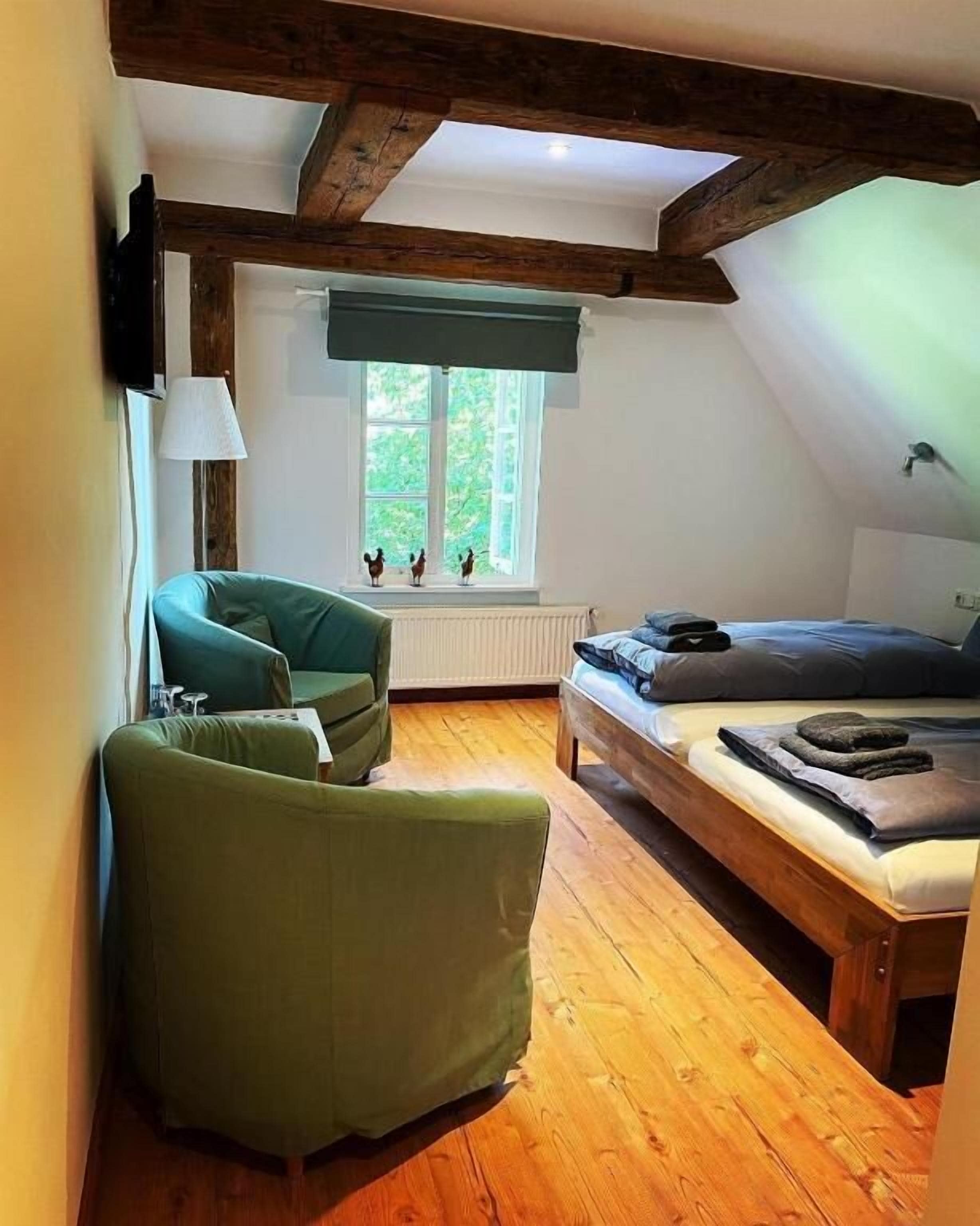 A cozy attic bedroom with a wooden ceiling, a bed with a blue mattress, a green armchair, and a window with a view of greenery.