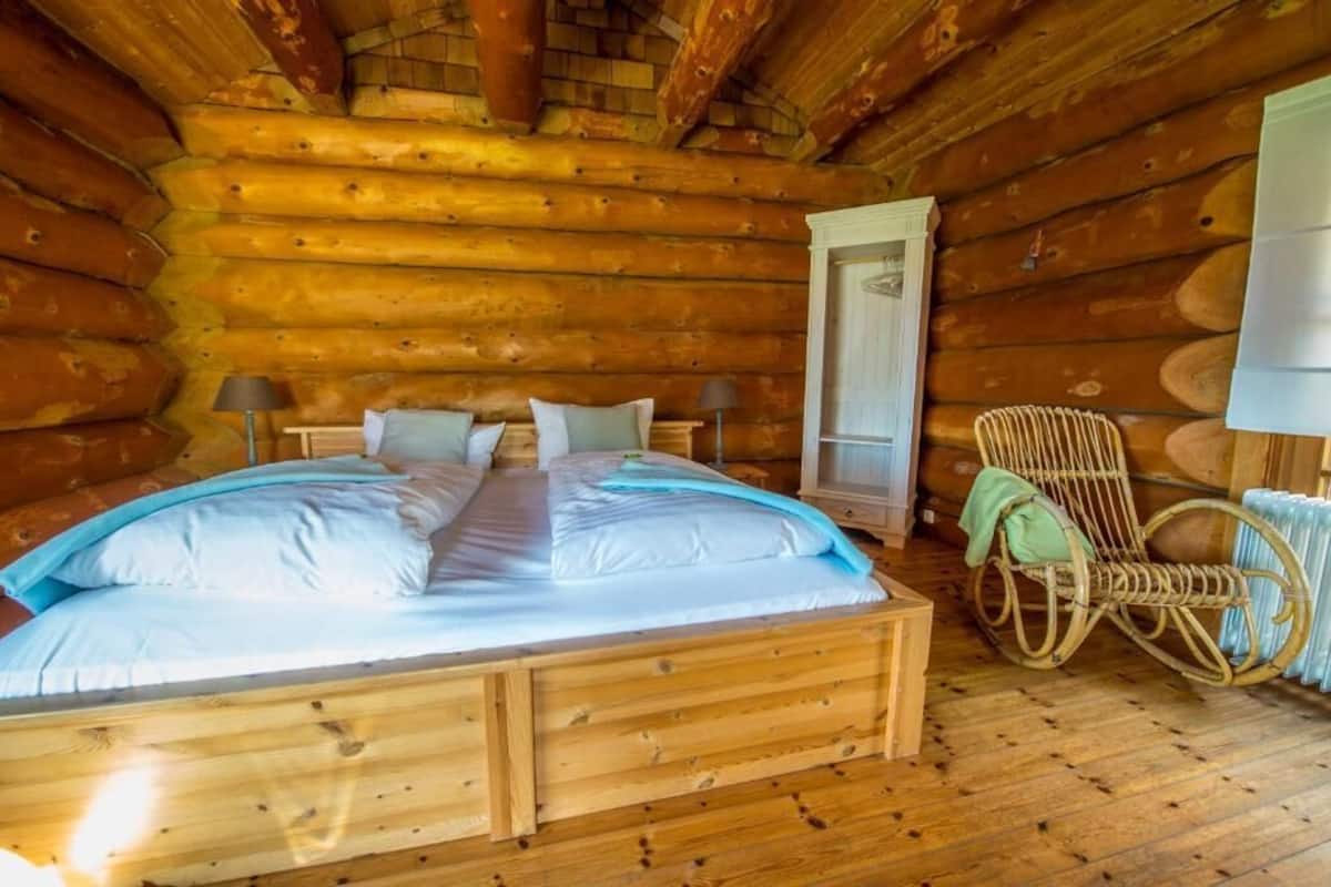 Un dormitorio de cabaña de madera con una cama de madera, dos mesitas de noche, una mecedora y una ventana.
