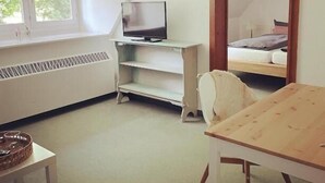 Ein Zimmer mit schrägem Dach, einem Fernseher auf einem weißen Regal, einem Bett mit Kopfteil, einem Holztisch und einem großen Spiegel.