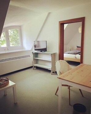 Een kamer met een schuine kap, een televisie op een witte plank, een bed met hoofdeinde, een houten tafel en een grote spiegel.