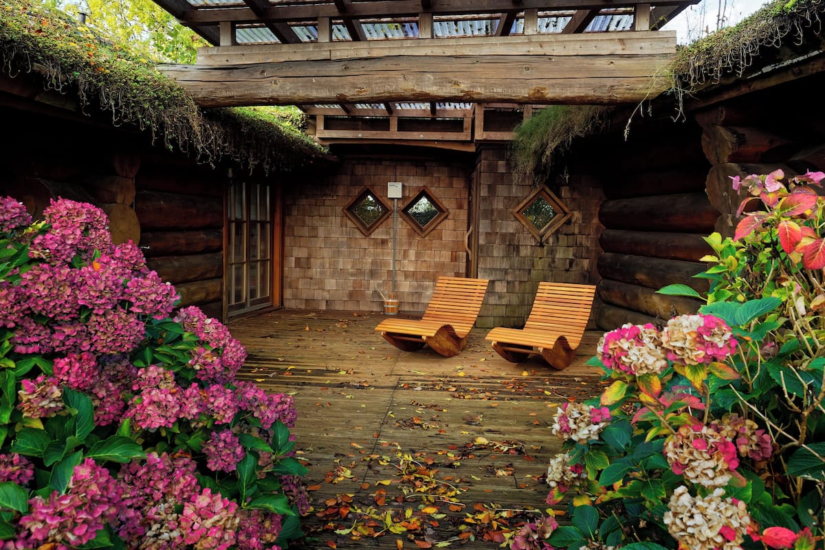 Una cabaña de troncos con terraza de madera, dos sillones de descanso y techo de paja.