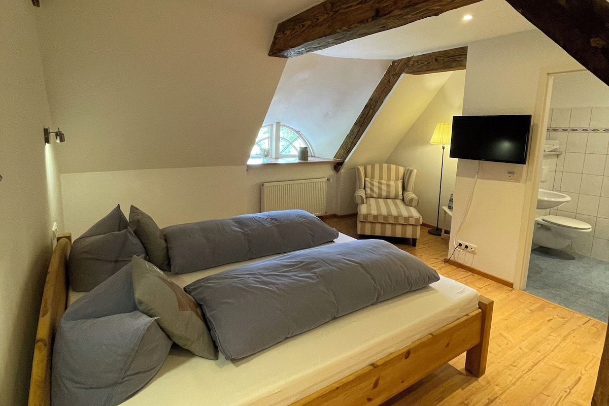 Un dormitorio con cama de madera, almohadas grises, una silla, un televisor y un baño visible a través de una puerta abierta.