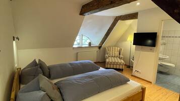 Un dormitorio con cama de madera, almohadas grises, una silla, un televisor y un baño visible a través de una puerta abierta.