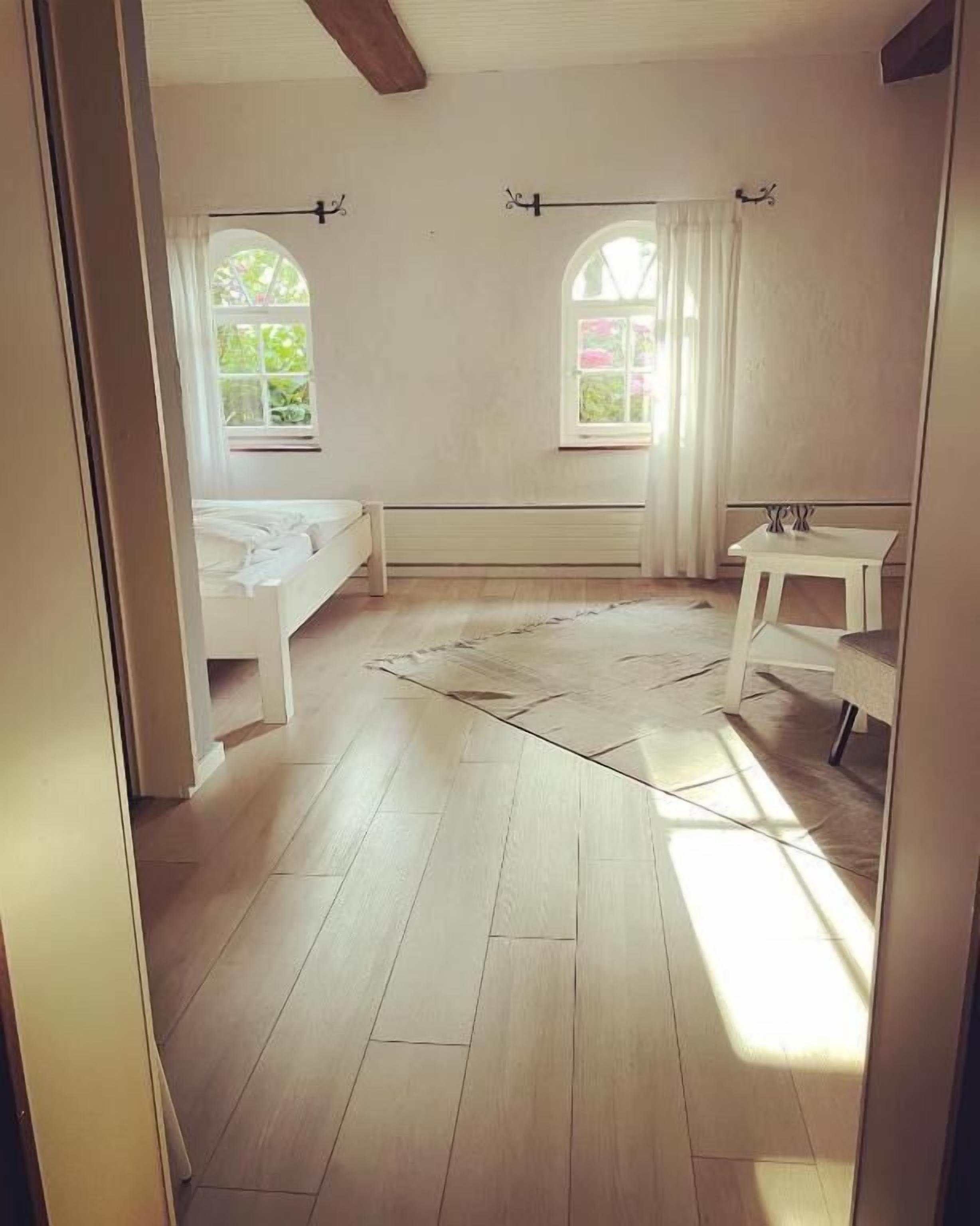 Ett rum med en säng, ett litet bord, två fönster med gardiner och ett element.