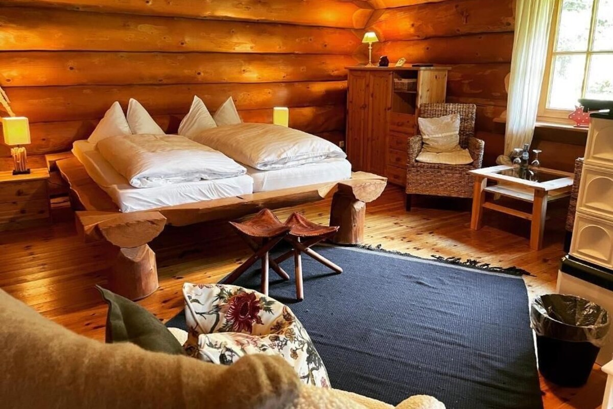 Ein Blockhauszimmer mit zwei Betten, einem Holztisch, einem Stuhl und einem Fenster mit Vorhängen.