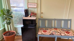 Un espace de vie compact comprenant un coin cuisine, un panneau décoratif représentant un bar à café, une plante en pot et un banc avec un revêtement floral.