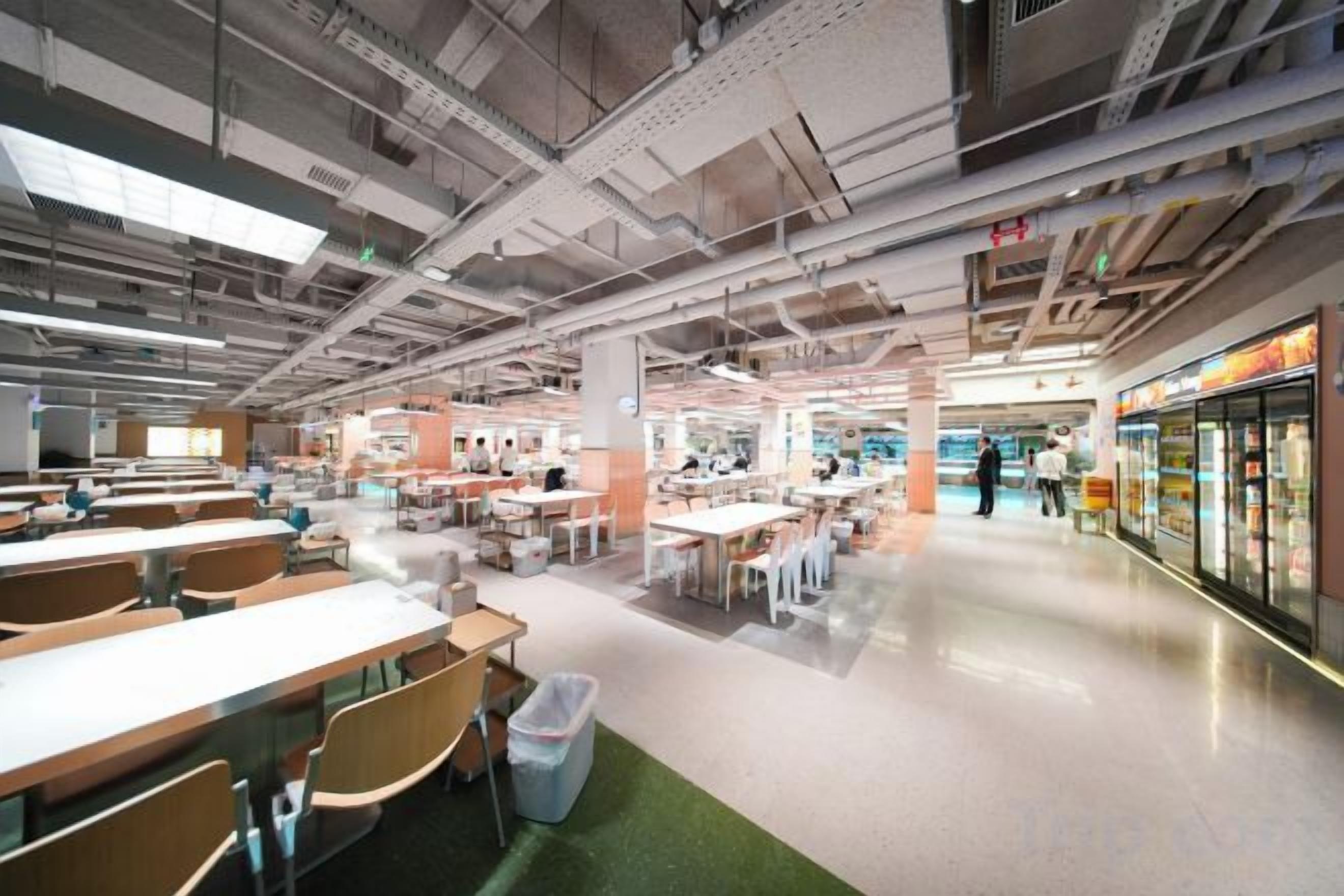 Uma cafeteria ampla com várias mesas e cadeiras, um balcão refrigerado para alimentos e um interior limpo e moderno.