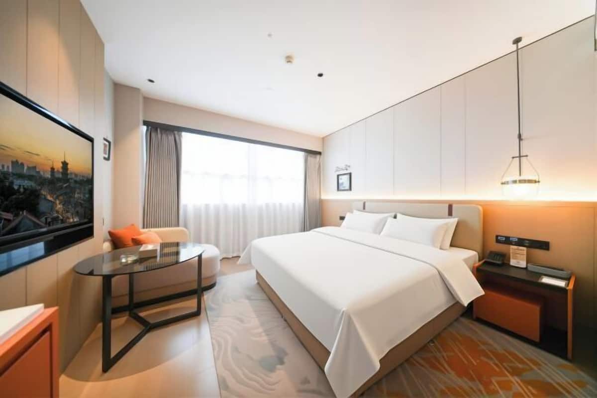 Ein Hotelzimmer mit einem Bett, einem Schreibtisch, einem Sessel, einem Fernseher, einem Fenster mit Vorhängen und einem Nachttisch mit Lampe.