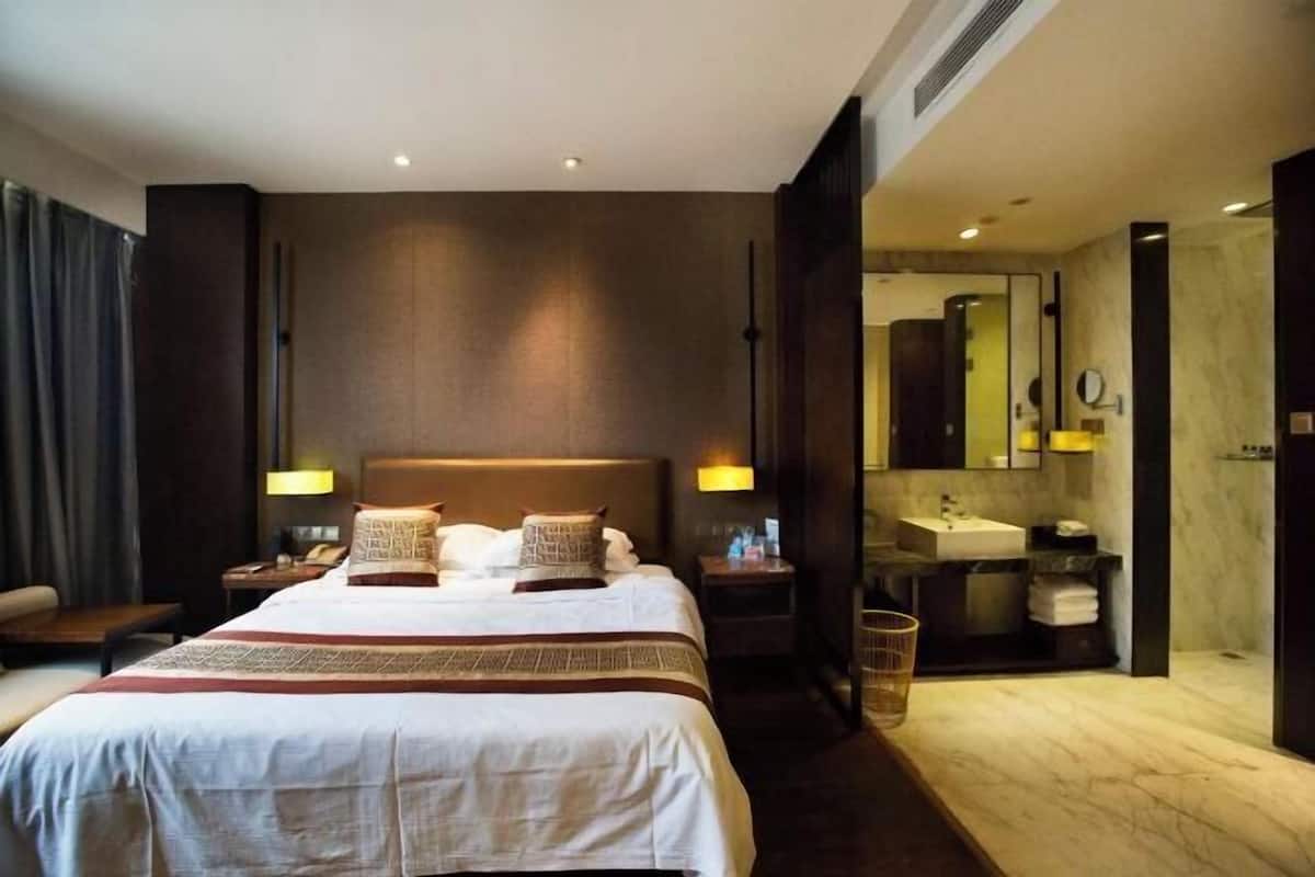 Ein Hotelzimmer mit einem großen Bett, Nachttischlampen, einem Nachttisch, einem Badezimmer mit Waschbecken und Spiegel sowie einem Mülleimer.