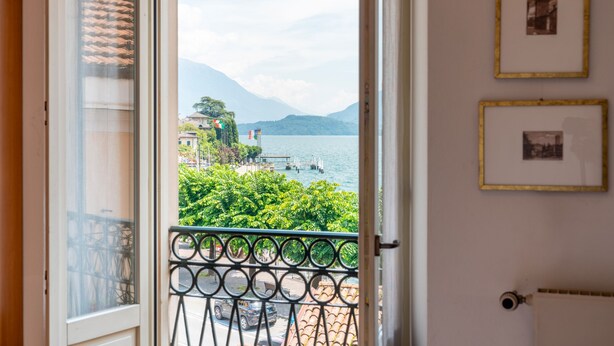 Un balcon doté d’une balustrade en métal, d’où l’on aperçoit un plan d’eau, une montagne au loin et un bâtiment surmonté d’un toit en tuiles.