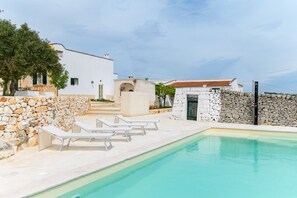 Un espace piscine doté de chaises longues blanches, de murs en pierre et d’un bâtiment blanc avec un toit recouvert de tuiles.