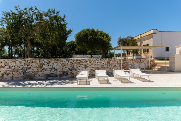 Un espace aménagé au bord de la piscine, avec des chaises longues, un mur en pierre et un bâtiment à la façade blanche.