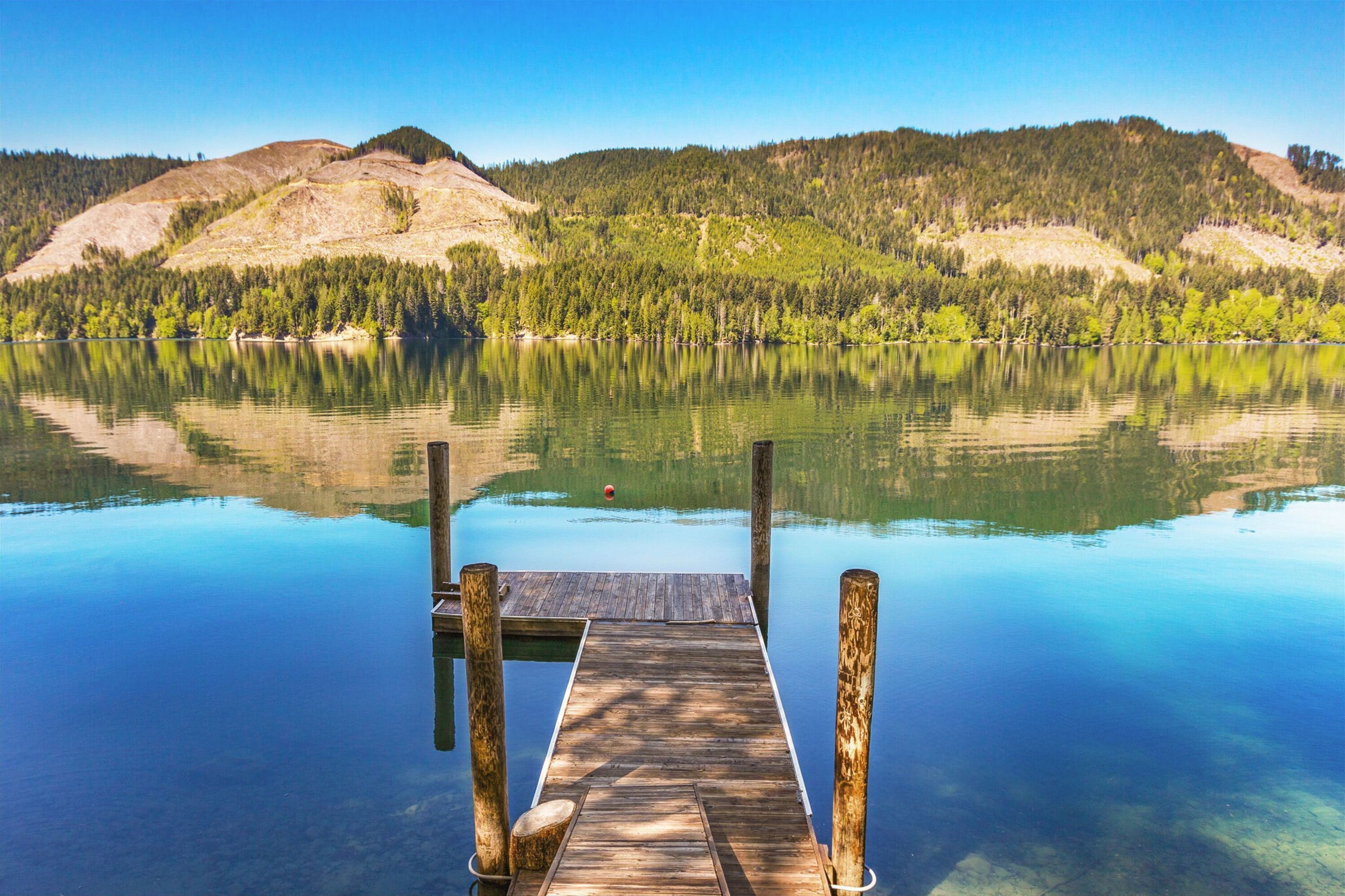 A wooden dock extending into a calm lake with clear reflections of surrounding hills and trees.