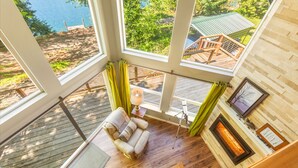 A room with large windows, a wooden deck, a sofa, a lamp, and a fireplace. - Yellow Tail Cabin in Hoodsport (Hoodsport)