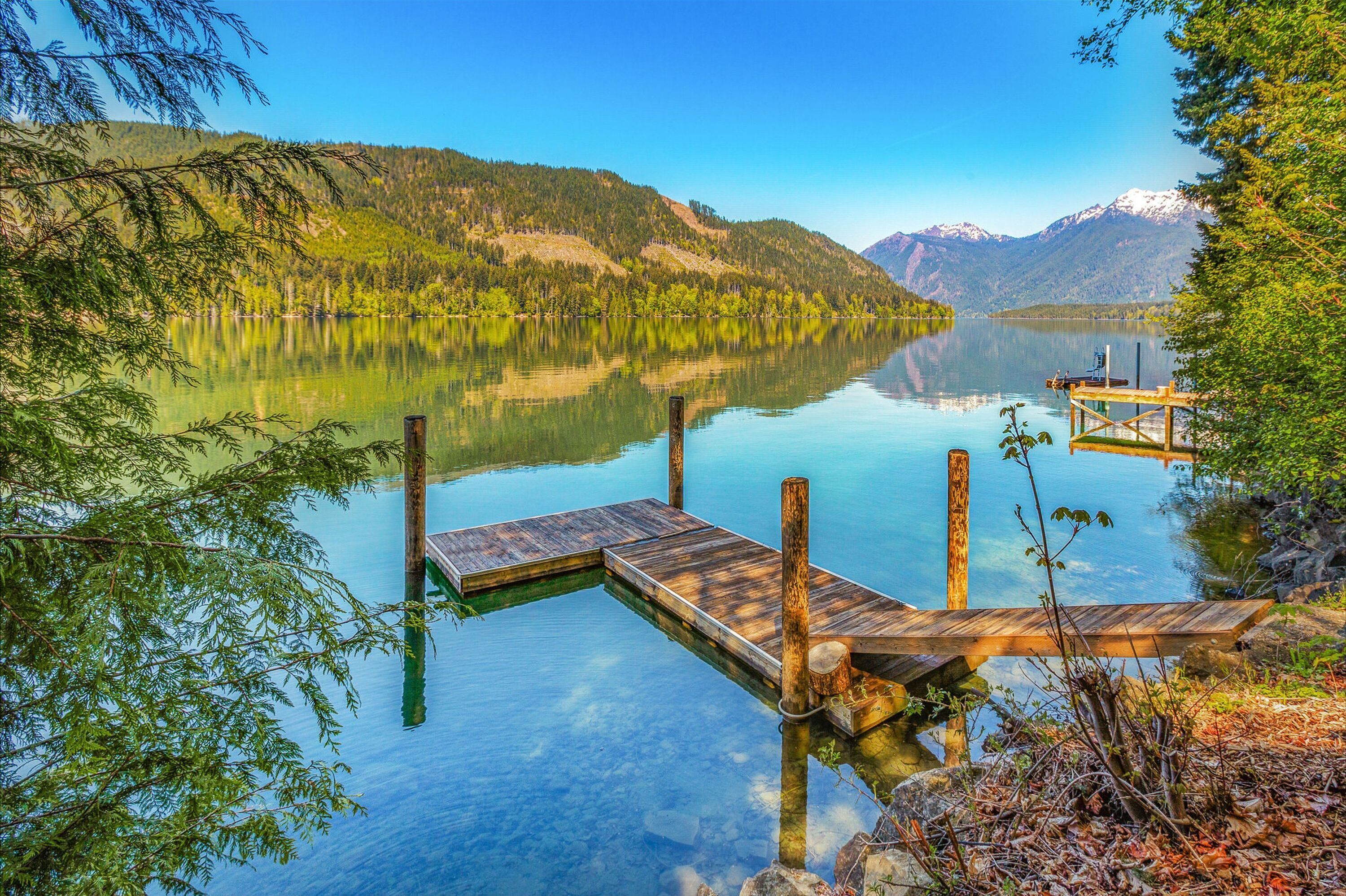 A wooden dock extending into a calm lake surrounded by forested hills and mountains.