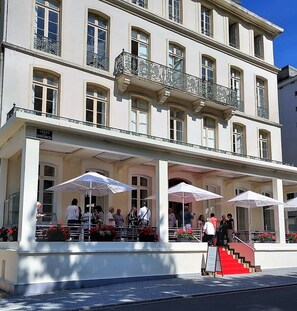 A multi-story building with a balcony, outdoor seating area with umbrellas, and a staircase leading to the entrance.