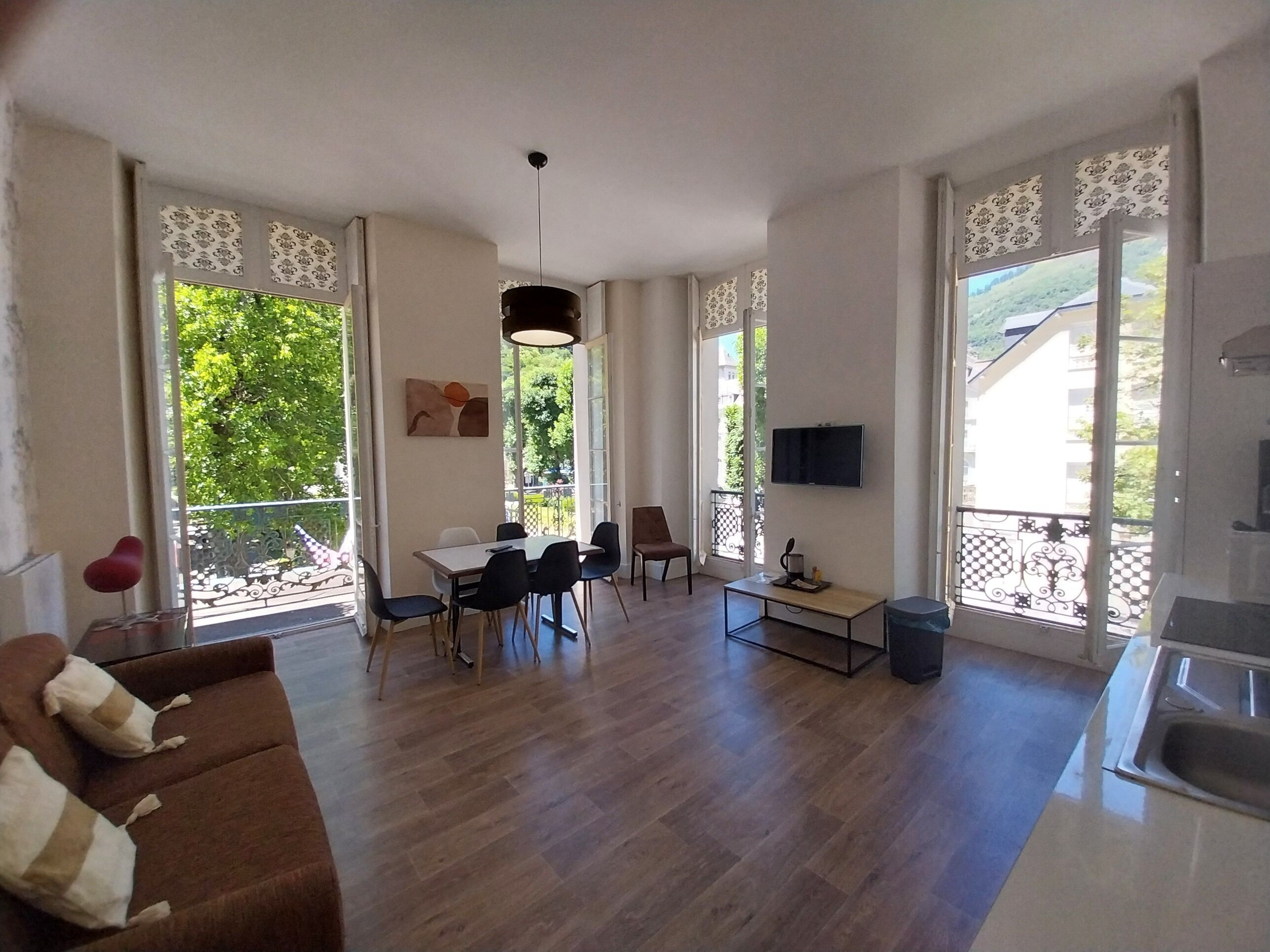Un vaste espace de vie comprenant un canapé, une table à manger, des chaises, un téléviseur fixé au mur et un balcon offrant une vue sur la verdure.