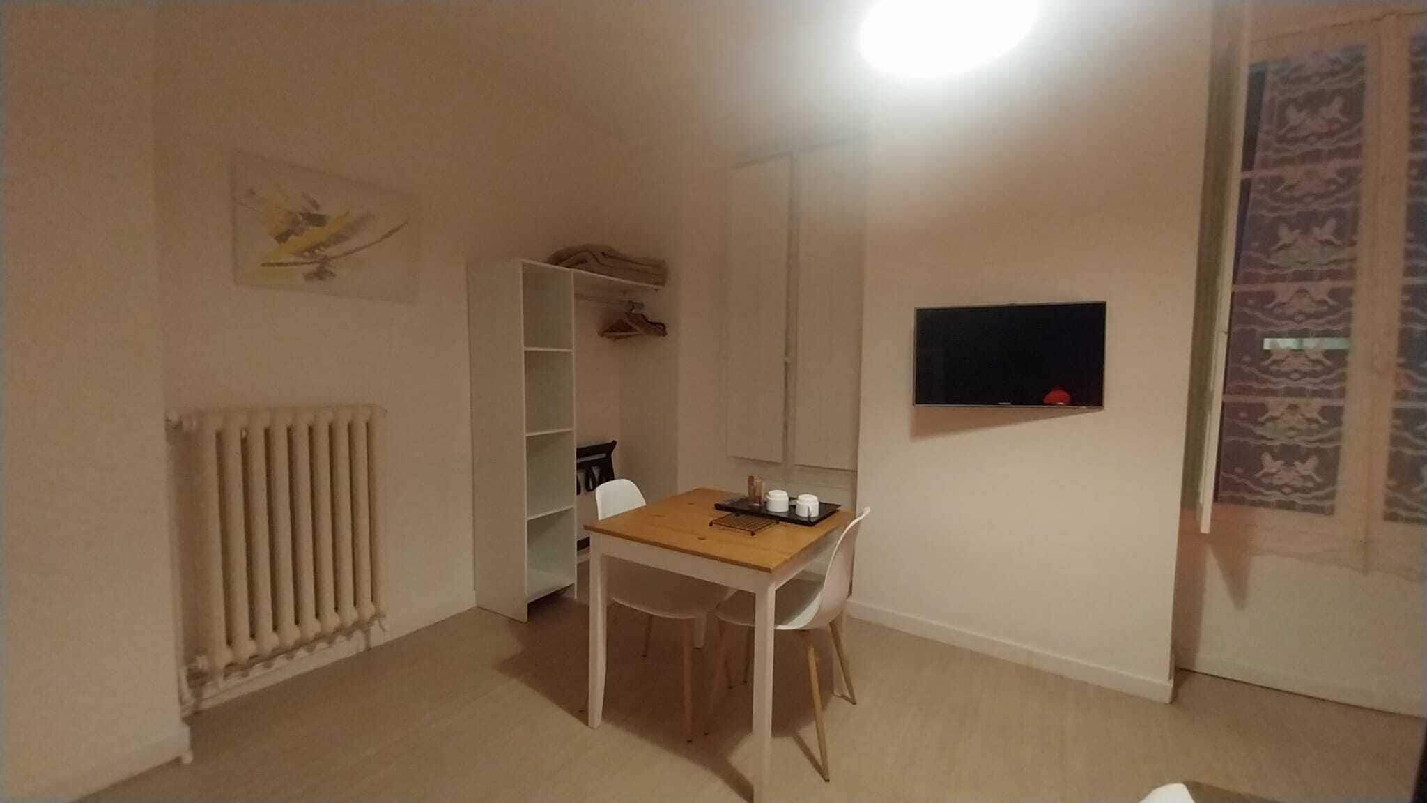 A room with a wooden table, white chairs, a radiator, a white wardrobe, and a mounted TV.