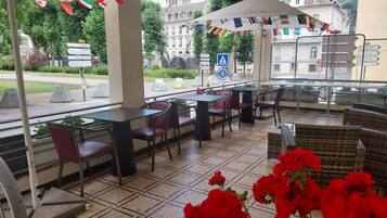 Un café en plein air avec des tables et des chaises, une vue sur un bâtiment en arrière-plan et une rue avec des panneaux de signalisation.
